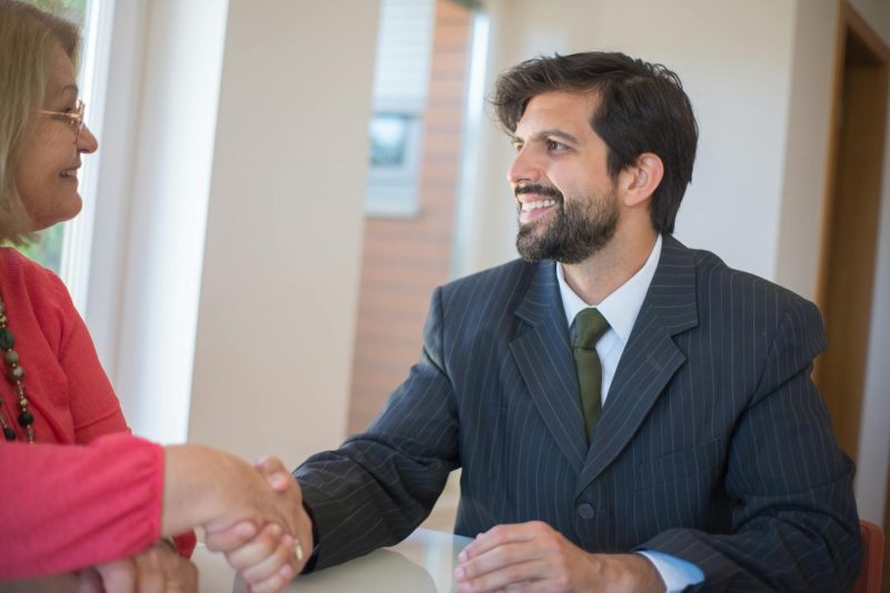 Professional real estate agent shakes hands with a senior client in a modern, well-lit indoor office setting.