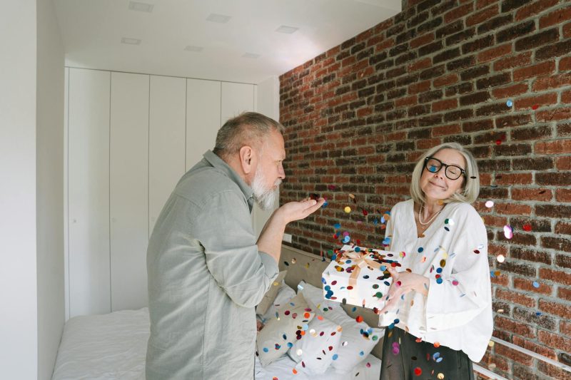 Happy senior couple celebrating indoors with confetti and a gift, surrounded by a cozy setting.