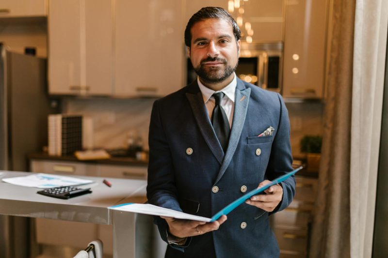 Confident businessman in a suit holding a folder, smiling in a modern office setting.