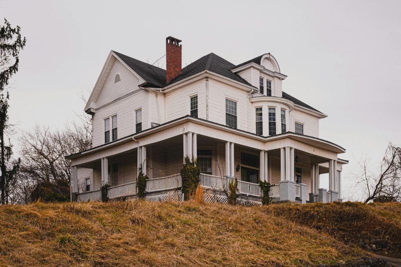 Historic white house with a wraparound porch surrounded by colorful autumn foliage.
